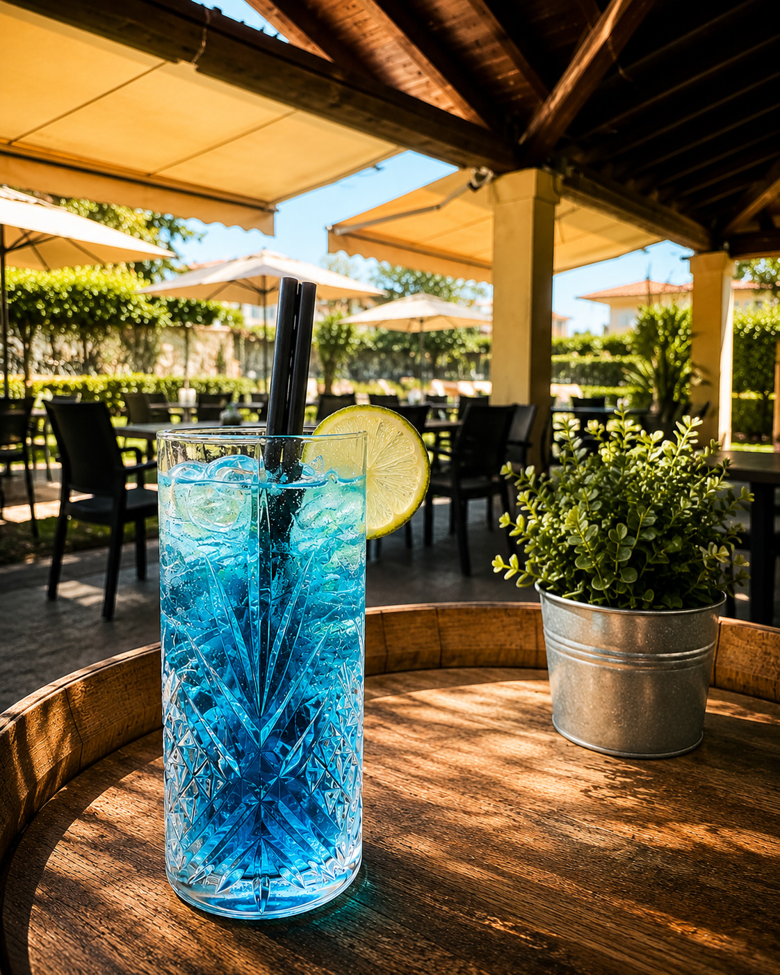 Cocktail ghiacciato azzurro con fetta di lime su un tavolo a botte, in un dehor soleggiato con ombrelloni e verde.